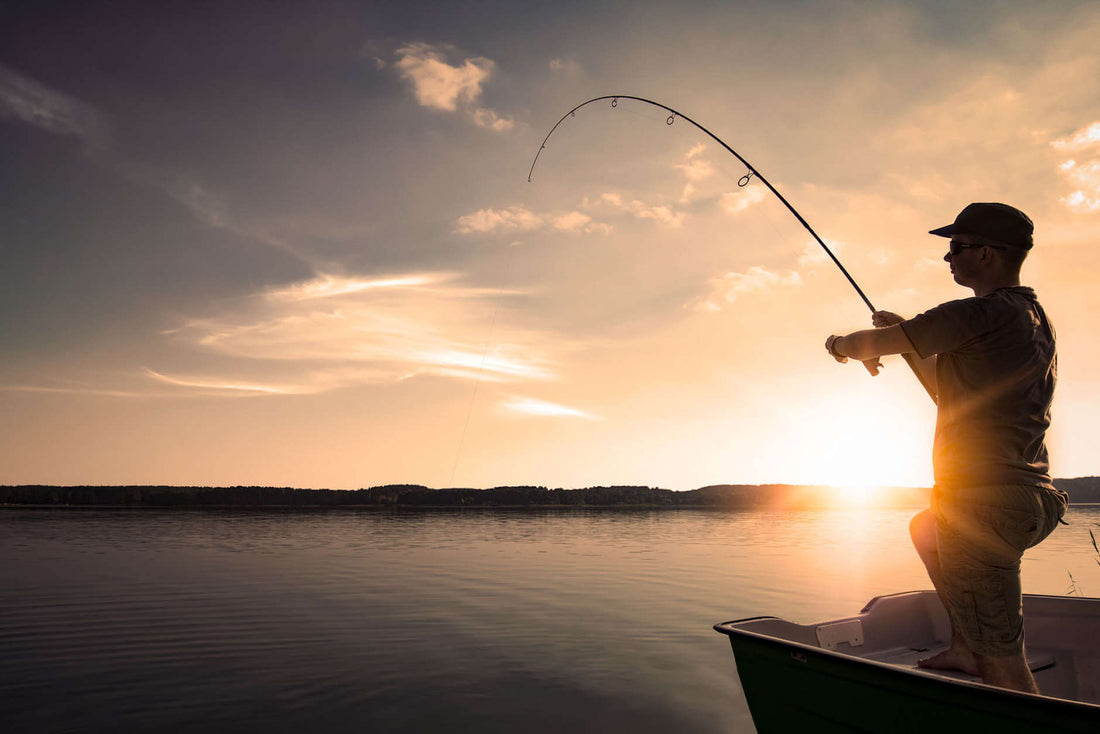 Silhouette of a fisherman casting a rod from a boat at sunset over a calm lake.